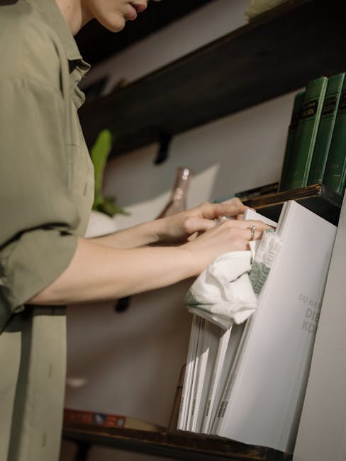 A woman wearing yellow cleaning gloves is carefully dusting a green leafy hanging plant in a modern indoor setting. She is facing a gray wall with painted white shelves that hold various cleaning supp