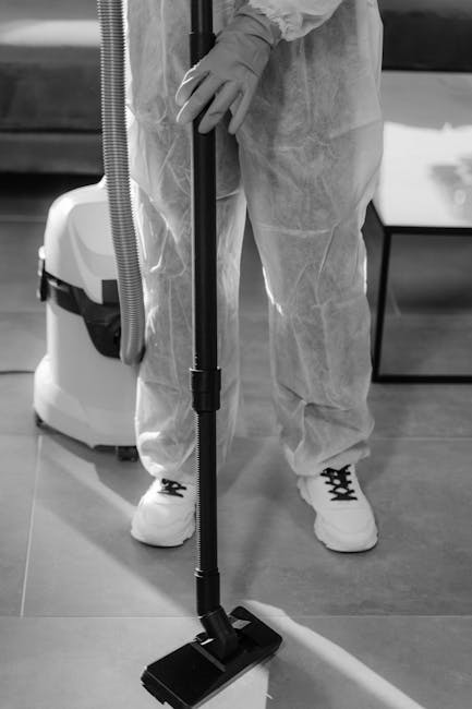 A person dressed in protective coveralls and gloves is using a black vacuum cleaner with a wide floor attachment to perform surface cleaning on a smooth, hard floor in an indoor room. The area appears