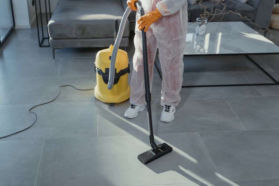 A person dressed in protective coveralls and gloves is using a black vacuum cleaner with a wide floor attachment to perform surface cleaning on a smooth, hard floor in an indoor room. The area appears